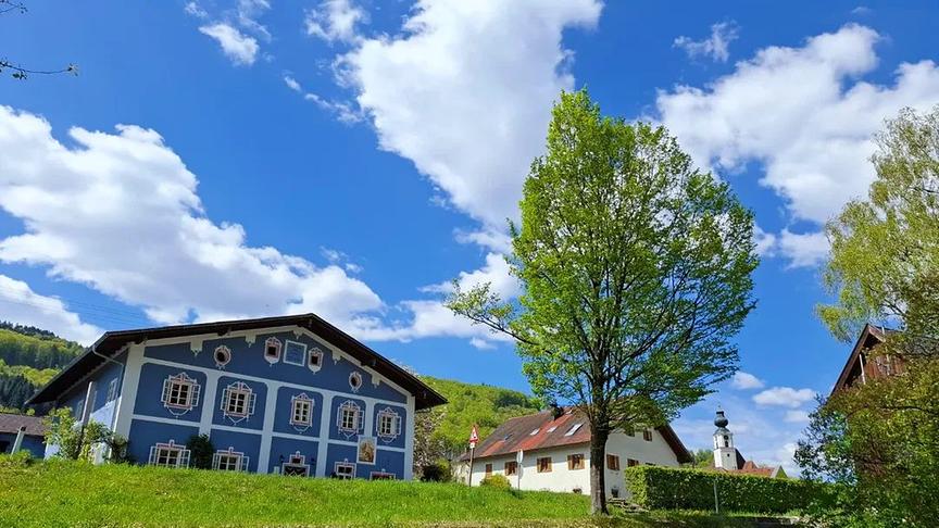 Mehrere Gebäude und eine Kriche umgeben von grünen Wiesen, Bäumen und Bergen unter Schönwetterhimmel.