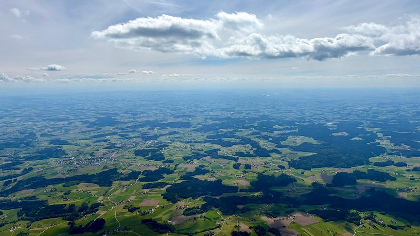 Weiter Blick über die Landschaft mit Feldern und Wäldern, die die Ebene fleckig wirken lassen. Darüber der Himmel mit Wolken, die Schatten auf die Landschaft darunter werfen.