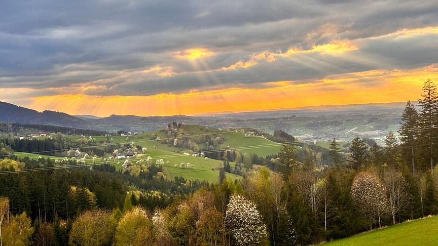 Blick über ein hügeliges Tal, durch die fast geschlossene Wolkendecke blitzen vereinzelt warm orange Sonnenstrahlen.