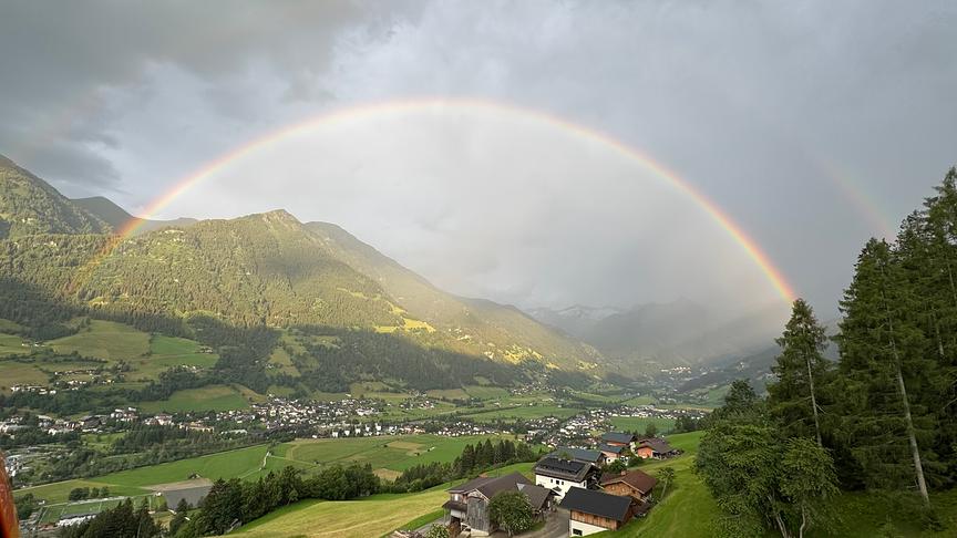 Ein vollständiger Regenbogen spannt sich von einem Berg zum anderen über ein bewohntes Tal. Sonnenschein fällt auf dem Regenbogen, während im Hintergrund noch dunkel und grau die Wolken in Schlieren hängen.