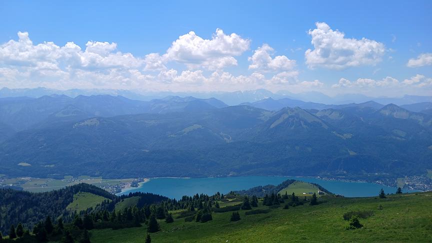 Blick hinab auf einen See umgeben von hohen Bergen, die sich immer weiter nach hinten im Hintergrund durchziehen. Am blauen Himmel ziehen Schönwetterwolken über die Landschaft.