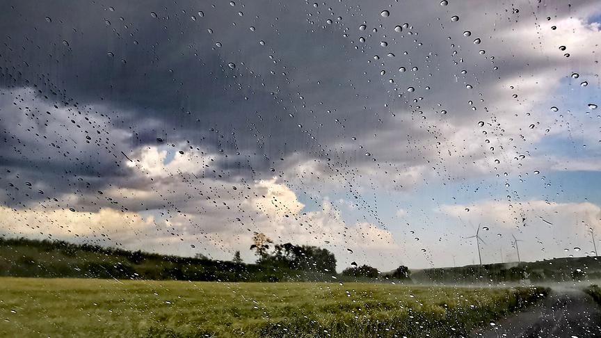 Regentropfen perlen an einem Fenster ab. Die Landschaft dahinter ist ein gelbes Kornfeld, Hügel und Wälder über denen sich dunkle Regenwolken bewegen.