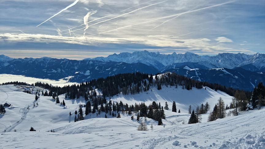 Verschneite Berglandschaft mit Spuren im Schnee, weiter unten gehen mehrere Personen, im Tal hält sich der Nebel