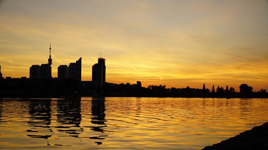 Sonnenuntergangsstimmung an einem Gewässer.Es dämmert bereits mit etwas oranglichem Restlicht des Sonnenuntergangs, das sich im Wasser spiegelt. Auf der anderen Seite des Gewässers sind die Silhouetten von mehreren hohen Gebäuder als Skyline zu sehen.