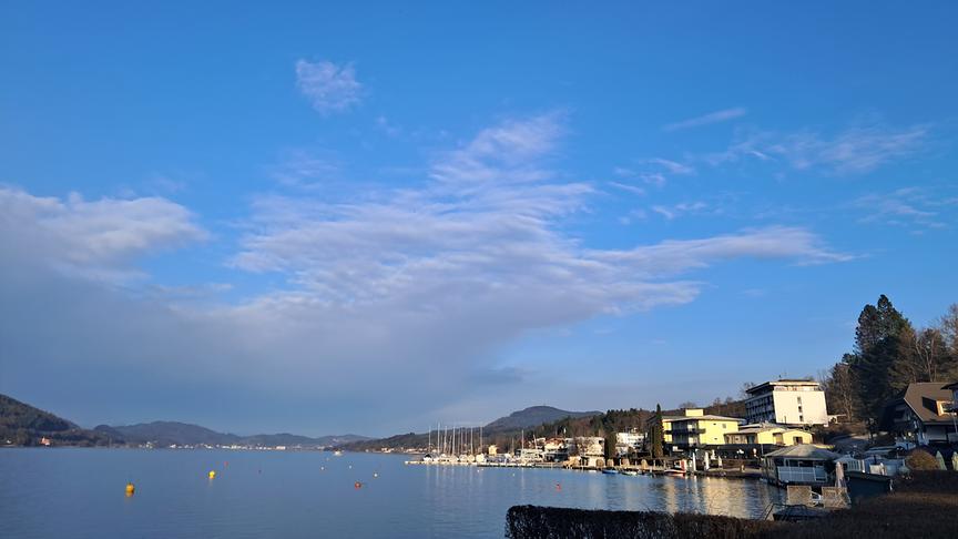 Über einem See zieht im Hintergrund eine graue Wolkendecke empor, im Vordergrund ist noch blauer Himmel und Sonnenschein