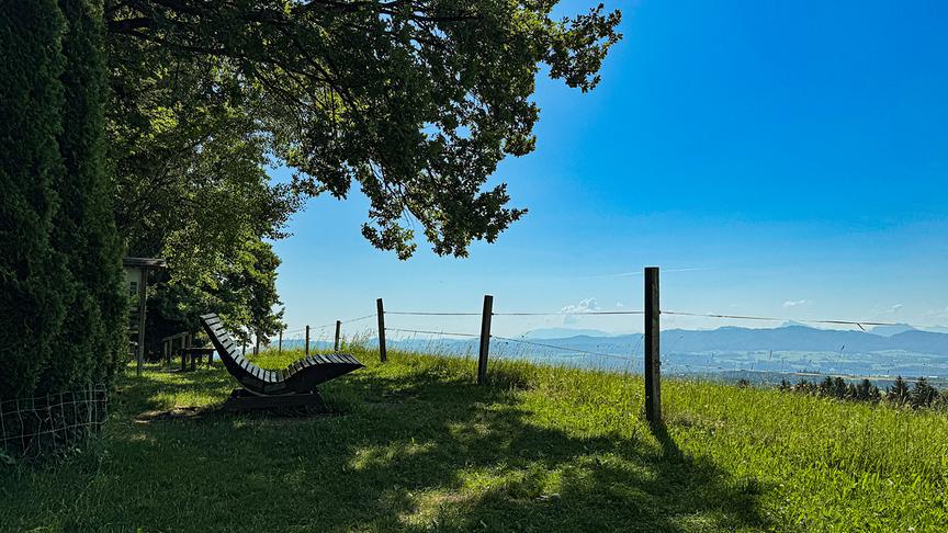 Eine Landschaftausnahme von einem durch Bäume schattigen Platz auf dem auch eine Liege steht. In Hintergrund sieht man hinunter auf ein Tal mit Bergen und darüber blauem, wolkenlosen Himmel.