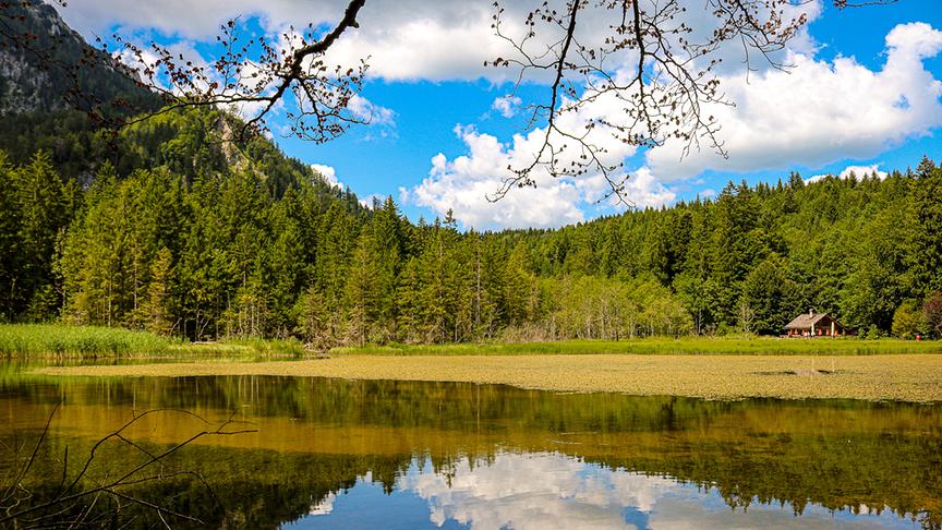 Ein dunkler See in dem sich die Berge und Bäume sowie der blaue Himmel mit Wolken spiegeln.