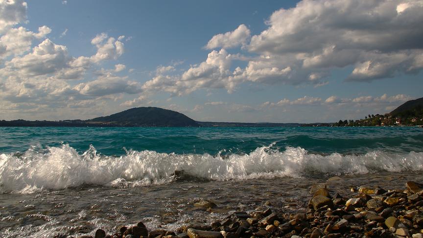 Kleine Wellen schlagen auf kiesiges Ufer. Dahinter erstreckt sich ein See bis zum anderen Ufer, wo sich ein paar Hügel erheben. Darüber blauer Himmel mit Wolken.