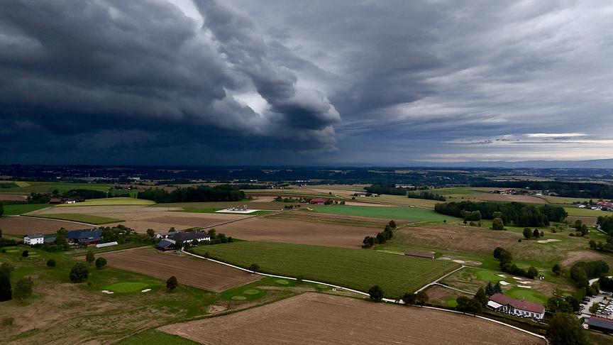 Luftaufnahme von Gewitterwolken, die sich über eine weite Landschaft mit Feldern, Bäumen und Häusern hinweg bewegt.