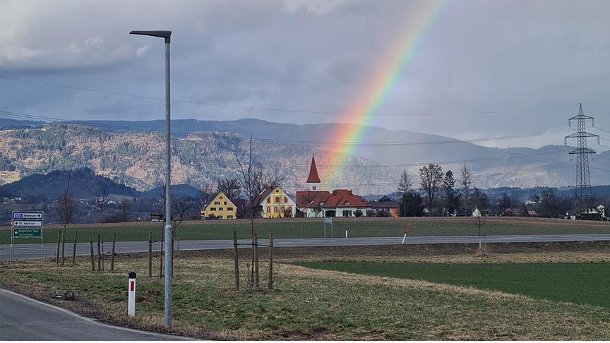 Der Regenbogen scheint genau auf die Kirche zu treffen, im Hintergrund Hügel und graue Wolken