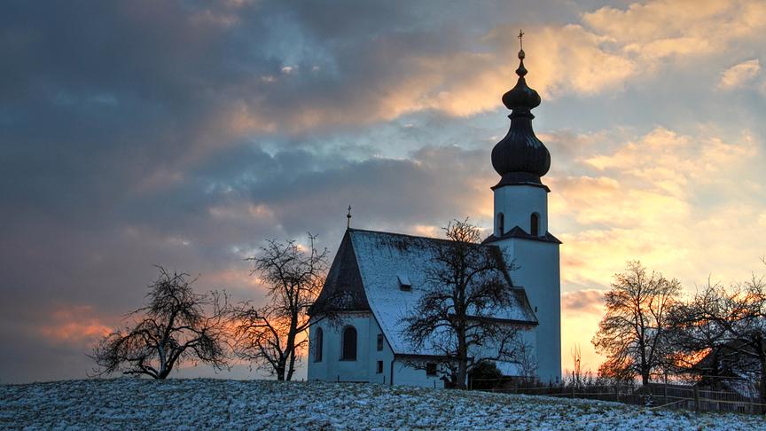 Kirche steht auf einem etwas verschneiten Hügel. Im Hintergrund färbt die Abendsonne die Wolken ein.