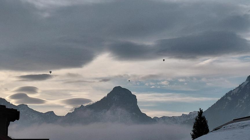Zwischen den emporragenden Bergen und der beeindruckenden, grauen Wolkenformation fliegen mehrere Heißluftballone.