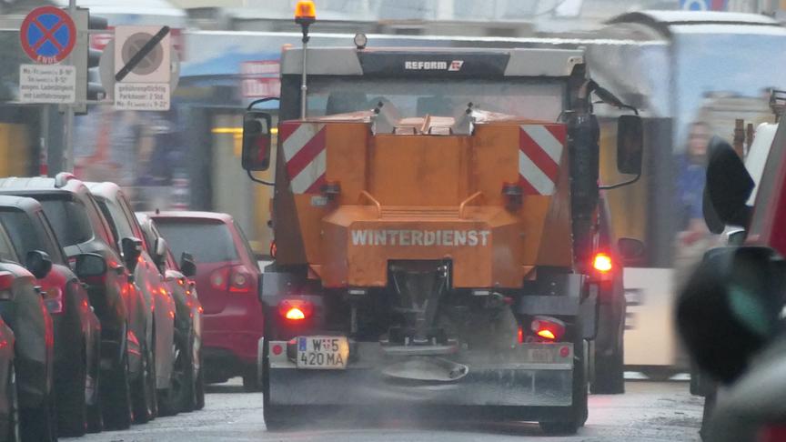 Winterdienst-Fahrzeug streut auf einer Straße mit Autos links und rechts während die Tram im Hintergrund vorbeifährt.
