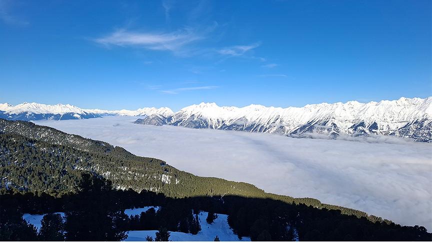 Zwischen den bewaldeten Bergen und dem gegenüberliegenden felsigen, verschneiten Bergpanorama hält sich die Nebeldecke im Tal