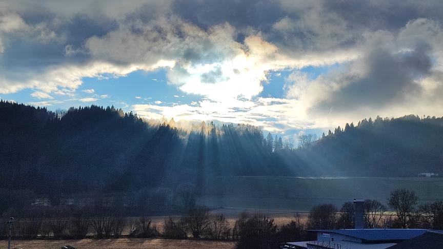 Sonne scheint durch die aufgebrochene Wolkendecke und über einen Wald hinweg