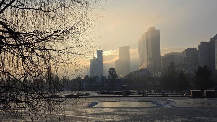 Hinter der teils noch zugefrorenen alten Donau stehen die Hochhäuser in vernebeltes Licht getaucht.