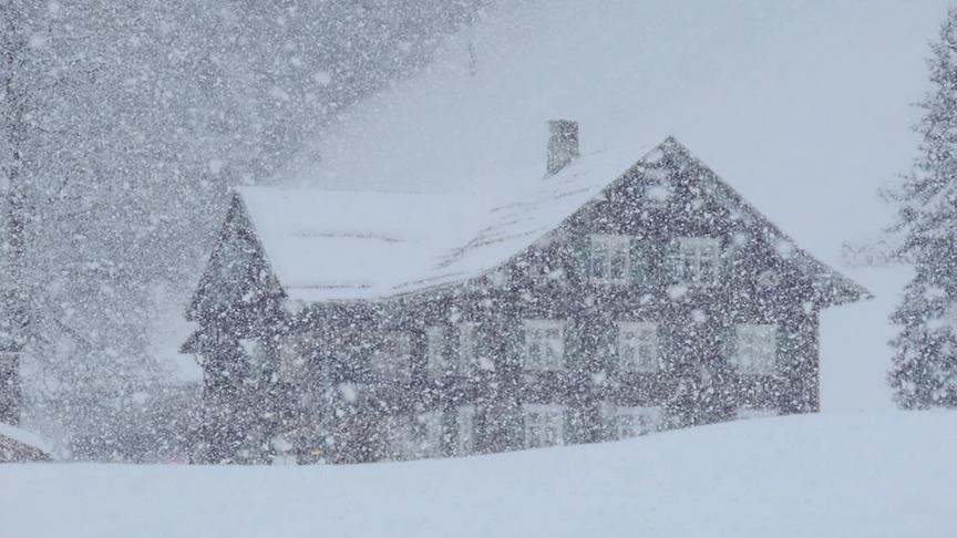 Ein dunkles Holzhaus, das nur unscharf durch den herrschenden starken Schneefall zu sehen ist.