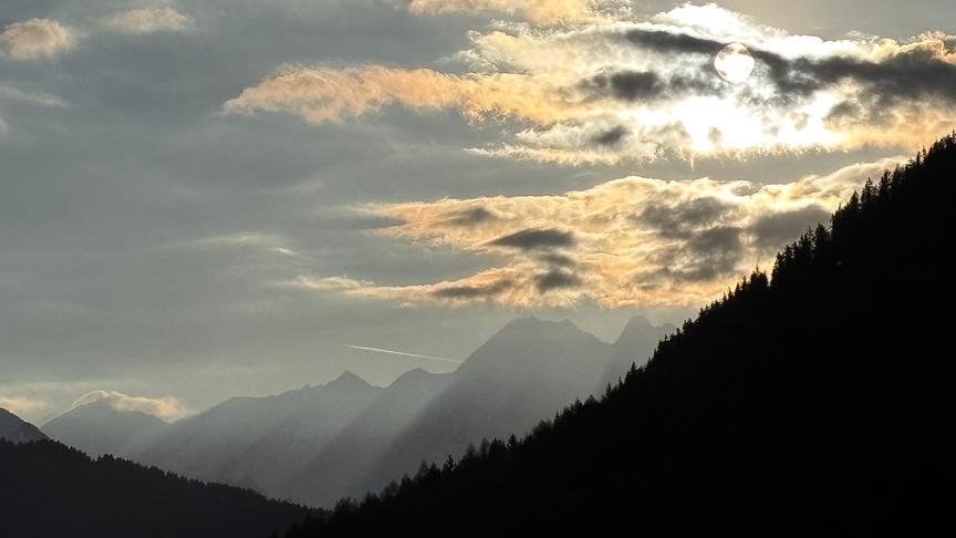Eine Kette von Bergen die shilouette-artig in milchiger Stimmung zu sehen sind. Die Sonne scheint durch Wolken.