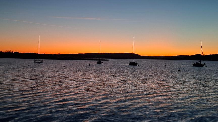 See mit ankernden Segelschiffen und der Silhouette von Bergen im Hintergrund in oranger Dämmerung.