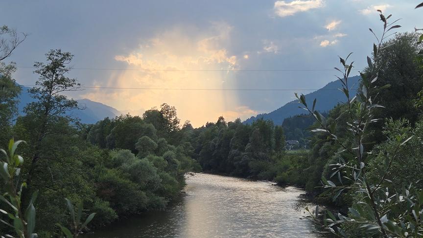 Ein Fluss schlängelt sich zwischen den dicht bewachsenen, bewaldeten Ufern hindurch. Im Hintergrund sind Berge zu sehen. Der Himmel ist fast vollständig von einer dunklen Wolkendecke bedeckt, die an einer Stelle jedoch aufgerissen ist und helle Sonnenstrahlen hindurchlässt.