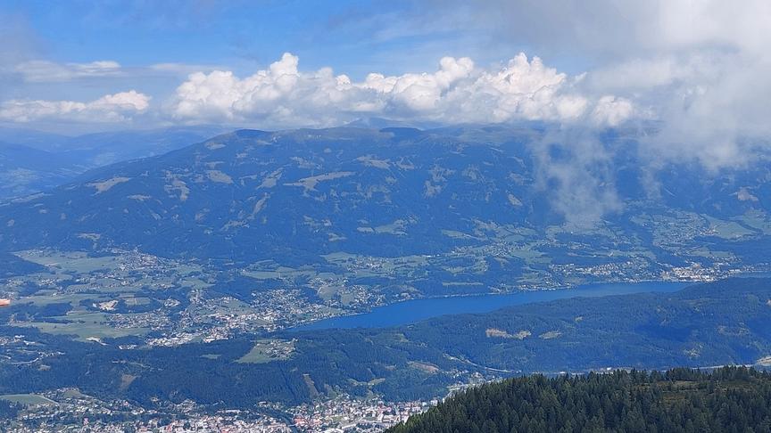 Weite Landschaft mit bewaldeten Hügeln und einem See im Tal mit Wolken darüber.