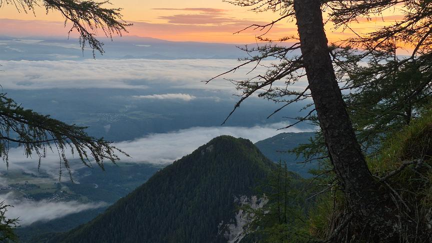 Von einem Berg herabfotografierte hügelige Landschaft mit Nebelschwaden bei Sonnenaufgang.