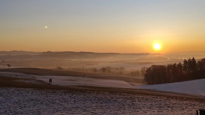 Über die leicht verschneite Hügellandschaft scheint die noch tiefstehende, aufgehende Sonne mit warmen Licht.
