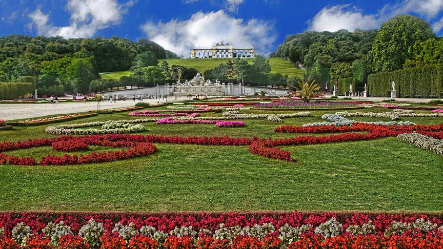 Gepflegte Parkanlage mit in Formen gepflanzten bunten Blumen, einem Brunnen im Hintergrund sowie der Gloriette auf dem Hügel darüber bei Schönwetter.