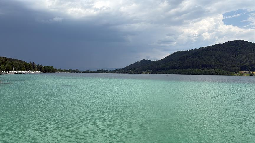 Türkis-farbener See mit Hügeln im Hintergrund. Am Himmel verdichten sich auf der einen Seite bereits dunkle Wolken.
