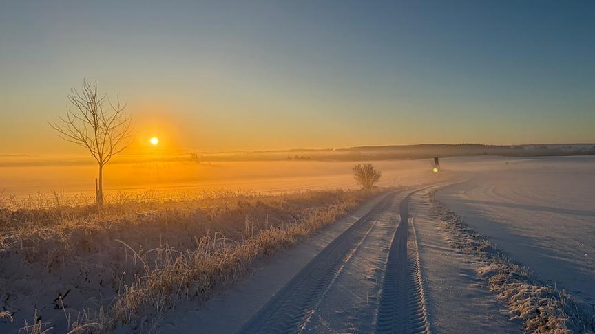 © Natalie Lunzer Ein verschneiter und eisiger Feldweg hinter dem die orangeleuchtende Sonne aufgeht