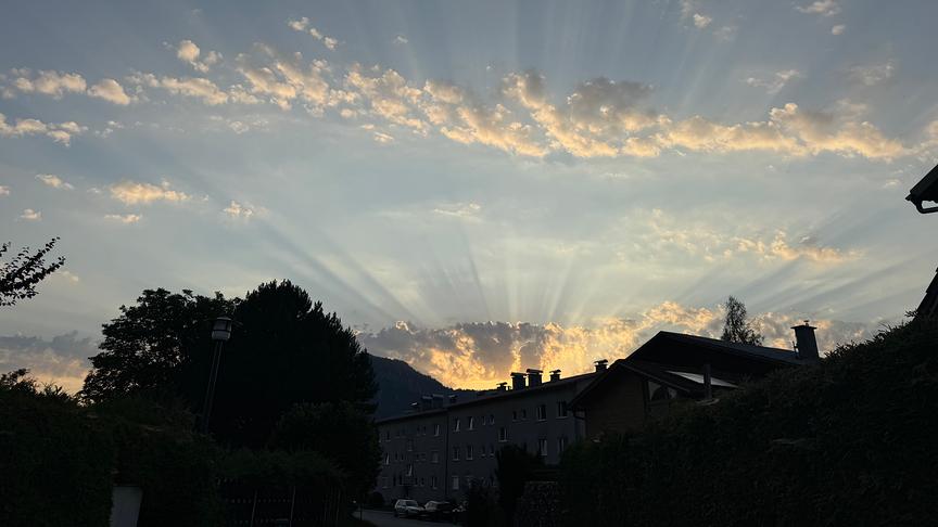 Stimmungsvolle Sonnenaufgangs-Aufnahme mit der Silhouette von Gebäuden und Bäumen im Vodergrund und einem Sonnenstrahlenbüschel im Hintergrund.