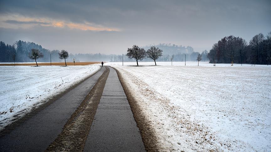 Leicht verschneite Landschaft in grauer, nebliger Stimmung mit einem Feldweg auf dem in der Ferne ein Spaziergänger geht.
