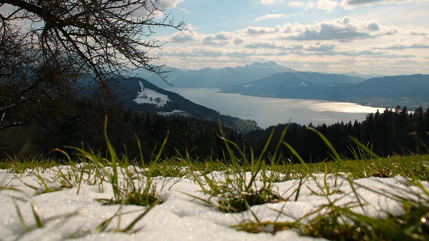 Im Vordergrund: Leicht verschneite Wiese auf einem Berg, im Hintergrund: Ein See umgeben von Bergen mit Wolken am Himmel