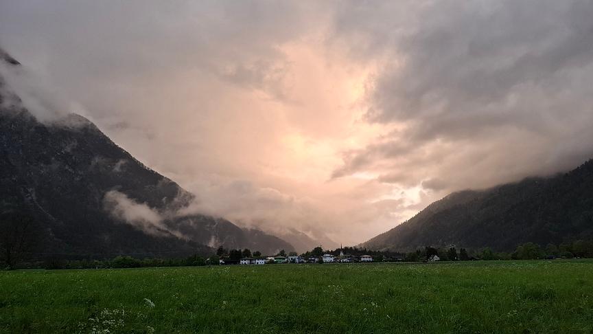 Tal mit einem Dorf, links und rechts steil abfallende Berge, dazwischen hängen tiefe, graue Wolken und etwas Licht erhellt sie.
