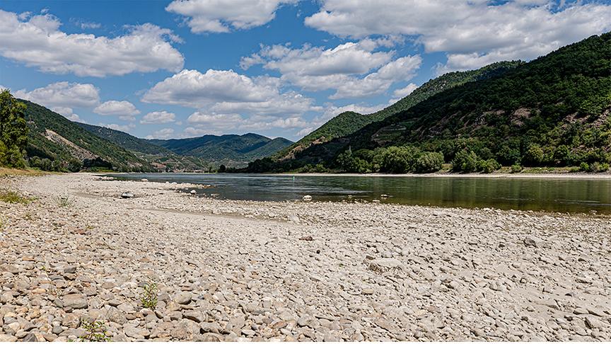 Die Donau führt wenig Wasser und gibt links und rechts breite, steinige Ufer frei. Sie windet sich durch sanfte, bewaldete Hügel unter wolkigem Schönwetterhimmel.