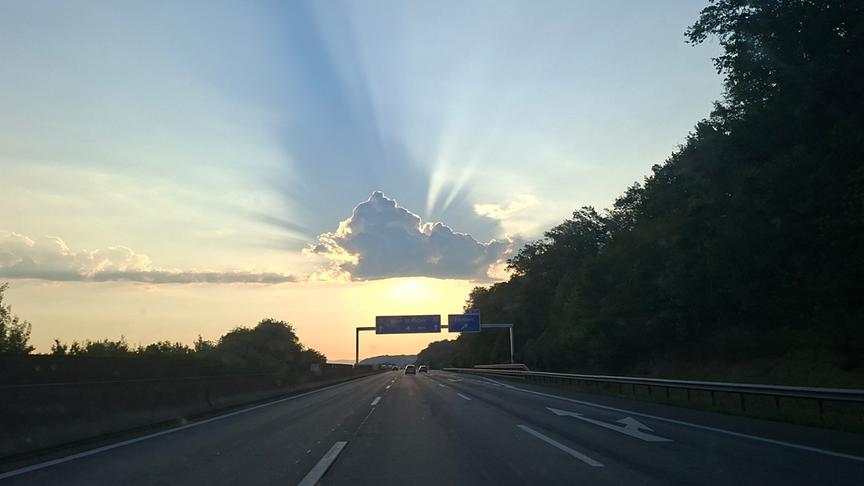 Geradlinige Autobahn über der mittig die Sonne hinter einer Wolke versteckt hervorscheint.