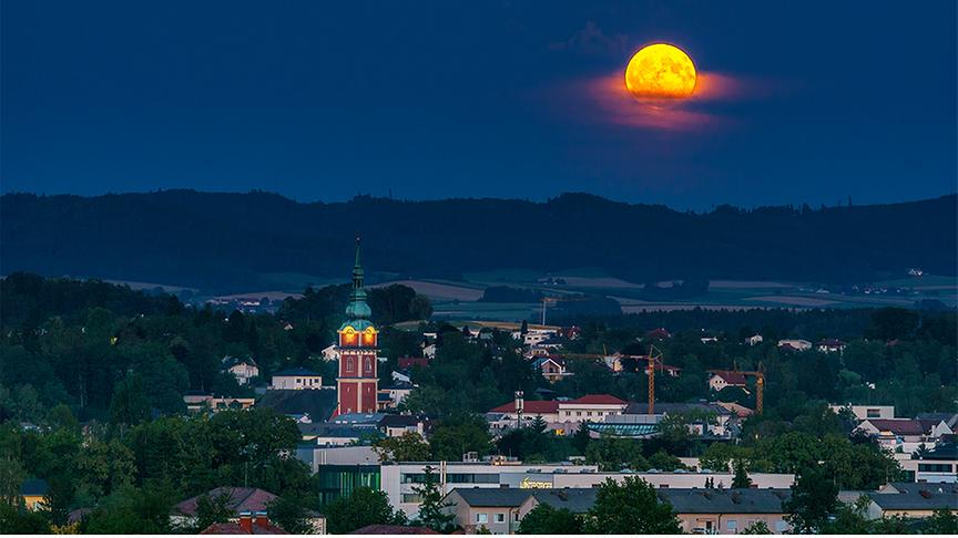Am dunklen Nachthimmel leuchtet der Vollmond warm-orange über einer Stadt umgeben von sanften Hügeln.
