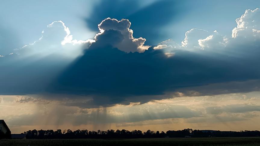 Eine große Wolke wird von hinten durch die Sonne bestrahlt. Die Sonnenstrahlen breiten sich vor allem über der Wolke aus. Unterhalb der Wolke ziehen sich Regenschleier über den Silhouetten von Bäumen zum Boden.