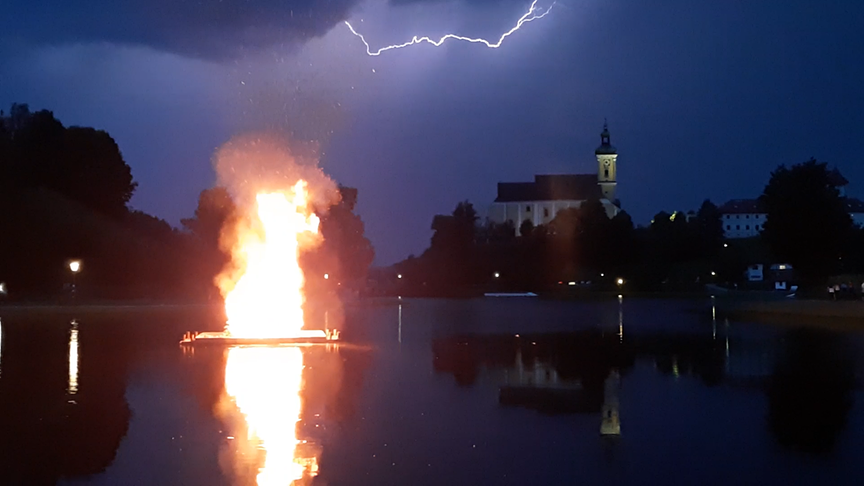 © Jasmin Kerschbaumer Auf dem Bild ist das schwimmende Sonnwendfeuer am Badesee, veranstaltet von den Oldtimerfreunden Waldhausen, bei einem Unwetter zu sehen. Im Hintergrund die Stiftskirche Waldhausen. (Jasmin Kerschbaumer)
