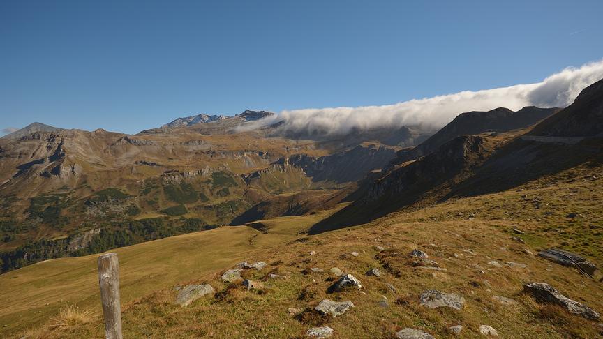 Weites Bergpanorama im Herbst, fotografiert von einem Berggipfel. Die umliegenden Hänge und Gipfel erstrecken sich in warmen Braun- und Erdtönen unter einem klaren Himmel.