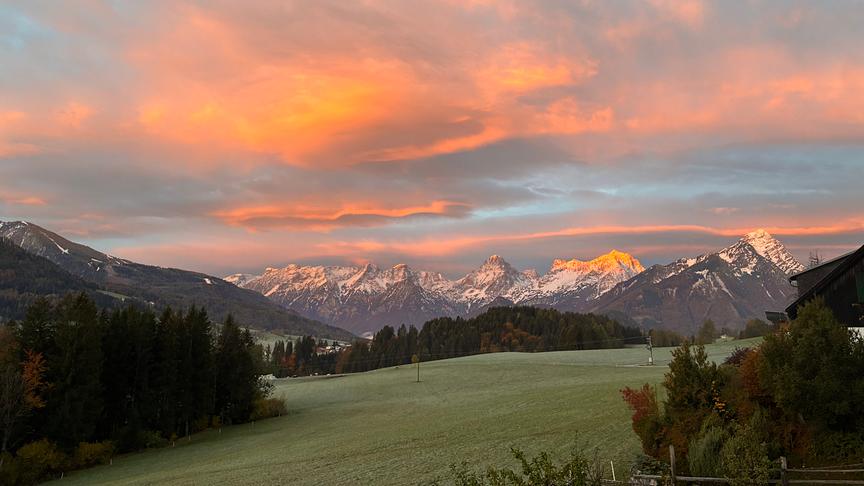 Bergpanorama mit verschneiten Gipfeln, grünen Wiesen und einem farbenprächtigen Abendhimmel.